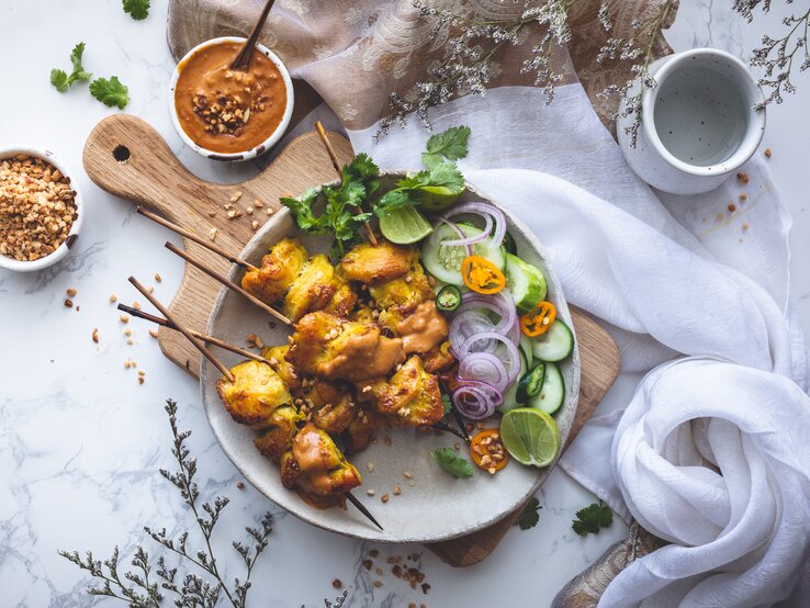 Ein Teller mit fünf Erdnuss-Satay-Spießen mit Salat, garniert mit Erdnussstücken, Koriander und zusätzlicher Erdnusssoße, die in einer kleinen Schüssel dazugereicht wird. Der Teller steht auf einem Holzschneidebrett, neben einem hellen Tuch vor einem weißen Hintergrund, von oben. 