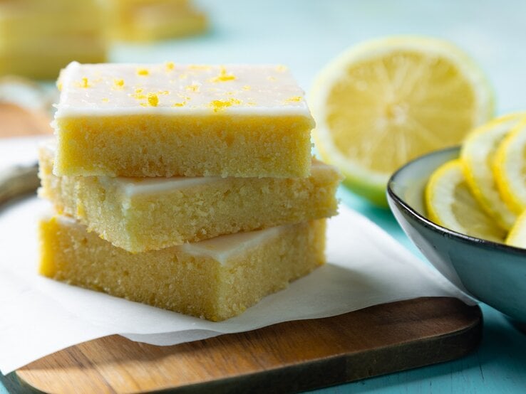 Drei Stücke Zitronen-Brownies gestapelt auf einem Brett, daneben eine Schale mit Zitronenscheiben und eine halbierte Zitrone.