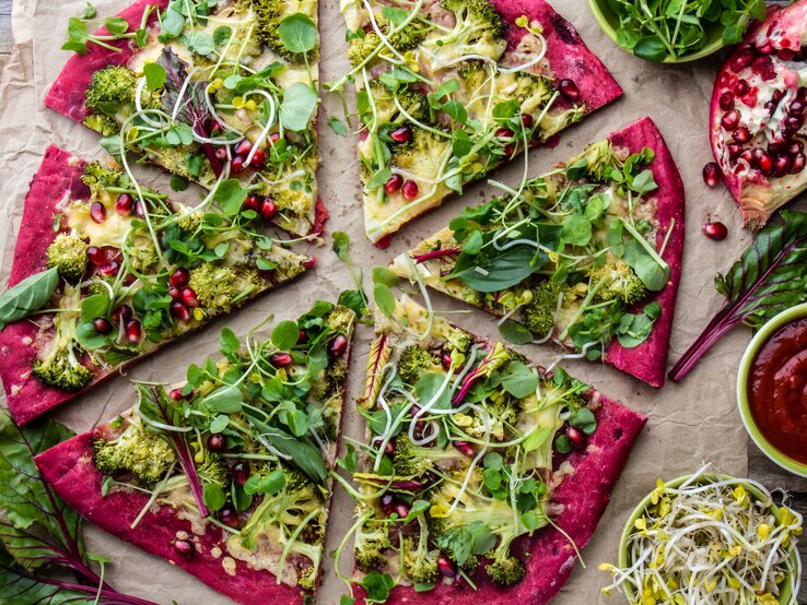 Eine in Stücke geschnittene Rotkohl-Pizza mit Salat, Sprossen und Granatapfelkernen in der Draufsicht.
