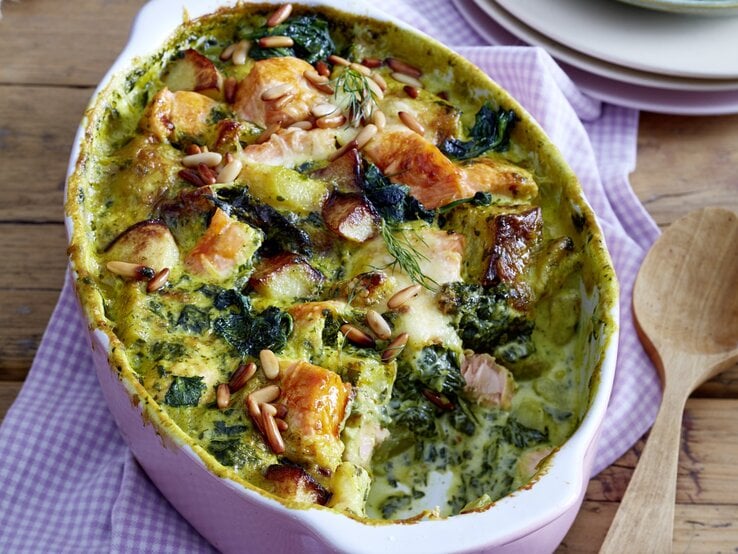 Kartoffel-Lachs-Auflauf mit Spinat in weißer Auflaufform auf blau-weiß-kariertem Küchentuch, darunter Holz, daneben ein Holzlöffel und im Hintergrund weißes Geschirr. 