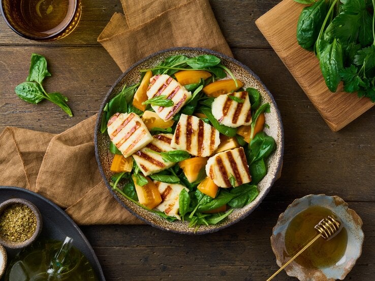 Untergund aus Holz. Darauf ein Teller  Gegrillter Halloumi-Salat mit orangen Tomaten. Drum herum liegen Spinatblätter und grobe Leinentücher