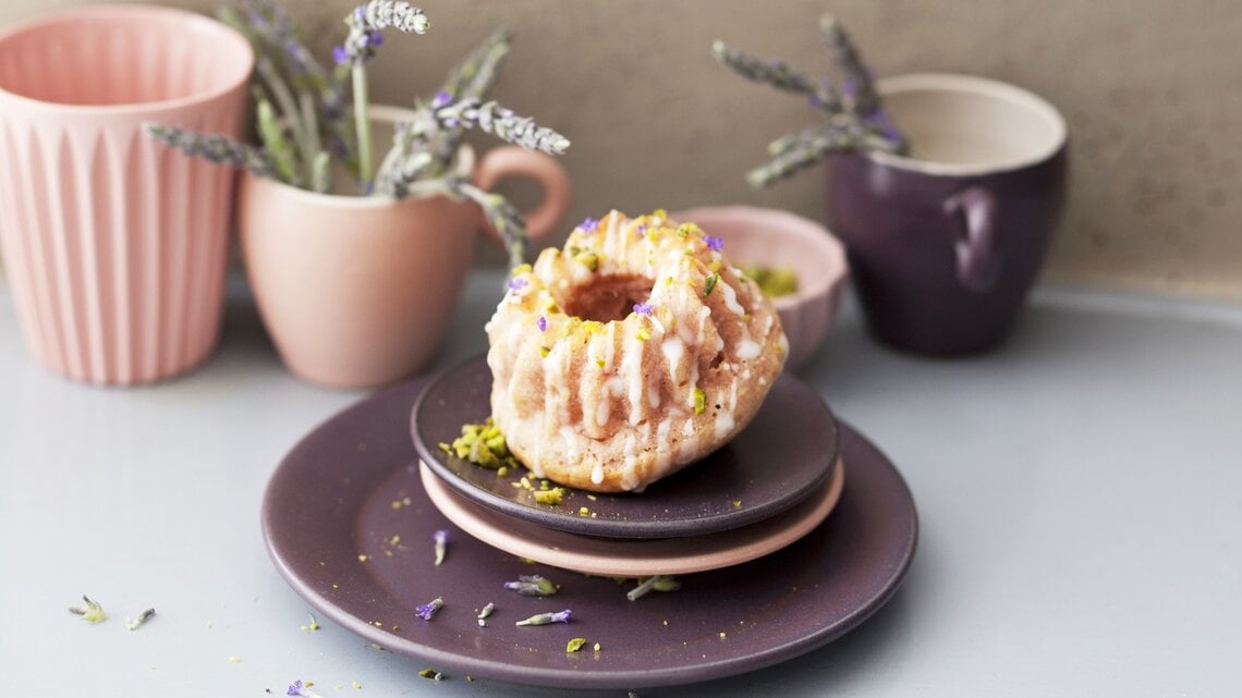 Ein Himbeer-Lavendel-Minigugelhupf auf Tellerstapel mit Geschirr und Lavendel im Hintergrund. Weißer Untergrund und beige Wand.