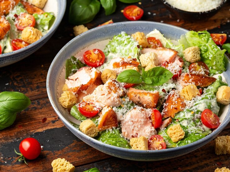 Zwei Teller mit dem Caesar Salat auf einem dunklen Holztisch. Drum herum Salat, Kräuter und Kirschtomaten.