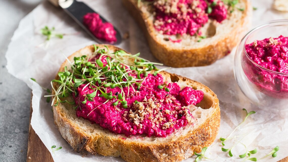 Ein paar Brotscheiben mit Pink Hummus, gehackten Nüssen und Kresse auf einem Holzbrett.