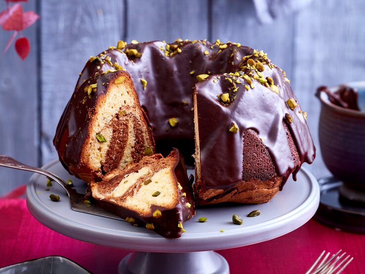 Ein angeschnittener Marmorkuchen auf einer Tortenplatte. Auf dem Tisch ist eine rote Tischdecke und dahinter eine rustikale Holzwand.