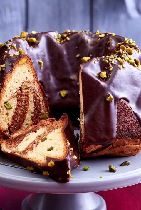 Ein angeschnittener Marmorkuchen auf einer Tortenplatte. Auf dem Tisch ist eine rote Tischdecke und dahinter eine rustikale Holzwand.