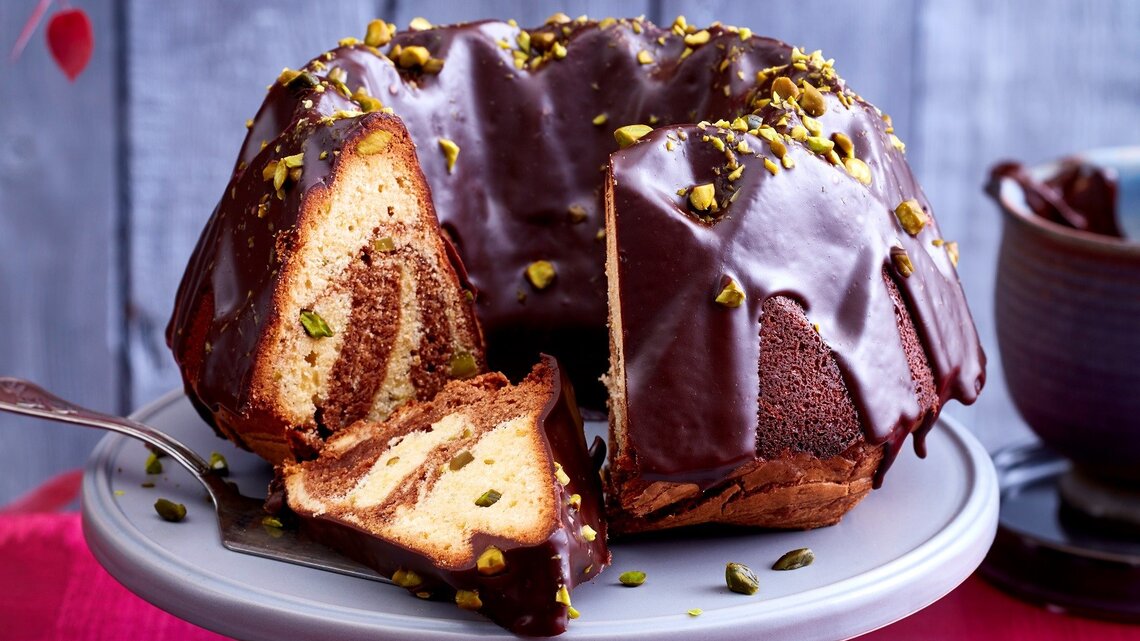 Ein angeschnittener Marmorkuchen auf einer Tortenplatte. Auf dem Tisch ist eine rote Tischdecke und dahinter eine rustikale Holzwand.