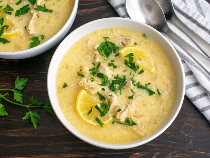 Draufsicht: Zwei Schüsseln mit der mediterranen Hühnersuppe Avgolemono stehen auf einem dunklen Holztisch. Die Suppe ist mit Petersilie und Zitronenscheiben garniert. Daneben liegen zwei Löffel und ein gestreiftes Küchenhandtuch.