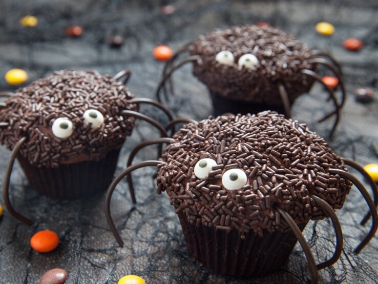 Süße Spinnen-Muffins mit Halloween auf dunklem Untergrund. Daneben liegen Schokolinsen in verschiedenen Farben. 