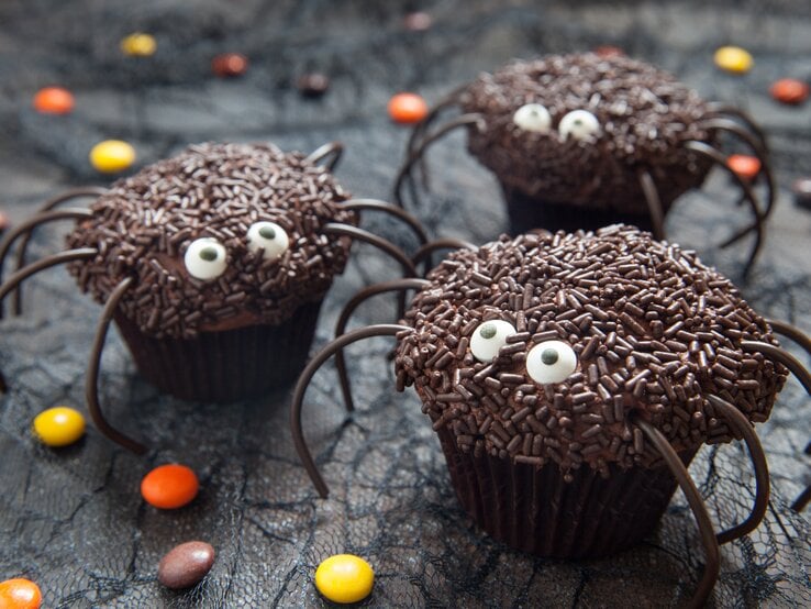Halloween-Muffins-Spinnen Süße Spinnen-Muffins mit Halloween auf dunklem Untergrund. Daneben liegen Schokolinsen in verschiedenen Farben.