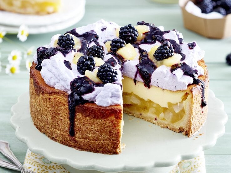 Auf einem Teller steht eine Birnen-Brombeer-Schmandtorte. Sie ist mit einer lila Creme verziert und mit Früchten bedeckt. Darüber wurde eine Brombeersoße gegeben. 