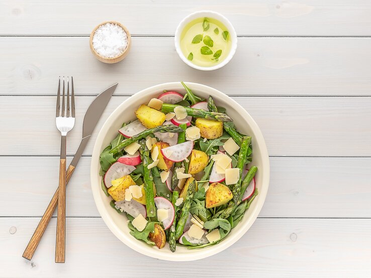 Eine Schale gegrillter Spargel-Kartoffel-Salat mit Radieschen und Rucola, daneben Besteck und eine schale Salz und Dip. Draufsicht.