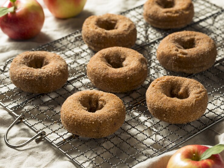 Apfelwein Donuts mit Zimt auf einem Gitter neben frischen Äpfeln auf einer hellen Tischdecke. 