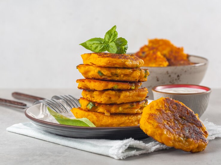 Mehrere Kürbispuffer auf einem dunklen Teller gestapelt mit einer kleinen Schale Gurkenjoghurt.
