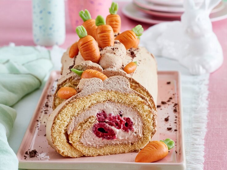 Auf einer Kuchenplatte ist eine Möhren-Biskuitrolle serviert. Sie ist mit Möhren aus Marzipan dekoriert. 