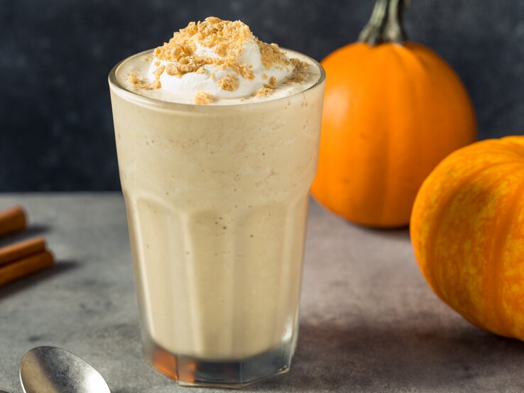 Frontalsicht: Ein Glas mit einem Kürbis-Milkshake mit Vanilleeis steht auf einem dunklen Untergrund. Neben dem Glas liegen Zimtstangen, eine Löffel sowie zwei kleine Kürbisse.