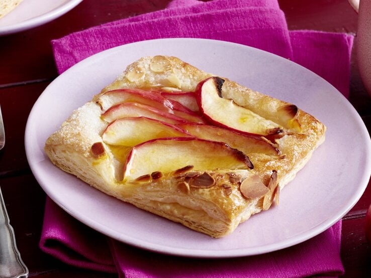 Auf einem lila Teller liegt ein Apfel-Knusper-Teilchen. Es ist goldbraun gebacken und sieht knusprig aus.
