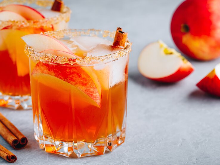 Schmeckt so schön fruchtig: unsere weihnachtliche Margarita. Zwei Gläser weihnachtliche Margarita mit Apfelspalten, Zimtstangen und Eiswürfeln. Daneben liegt ein angeschnittener Apfel.