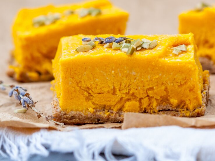 Drei Stücke von dem Kokos-Kürbis-Cheesecake auf Backpapier und einem grauen Tisch. Sehr nah von der Seite fotografiert.