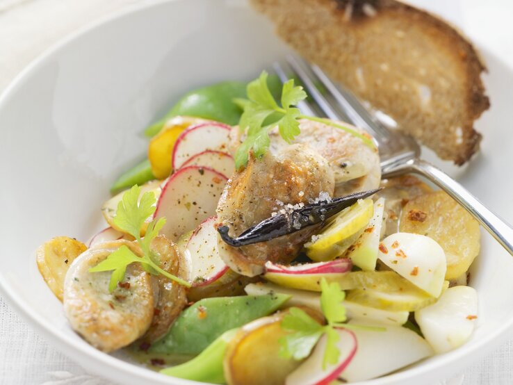 Bratwurstsalat-Thüringer-Art Ein weißer Tiefer Teller mit dem Bratwustsalat auf einem Tisch mit weißer Tischdecke sehr nah von schräg oben fotografiert.