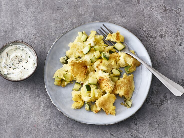 Halloumi-Zucchini-Schmarrn Ein heller Teller mit Halloumi-Zucchini-Schmarrn und einer Gabel steht auf einem grauen Untergrund. Links neben dem Teller steht eine kleine Schmale mit Schmarrn-Dip.