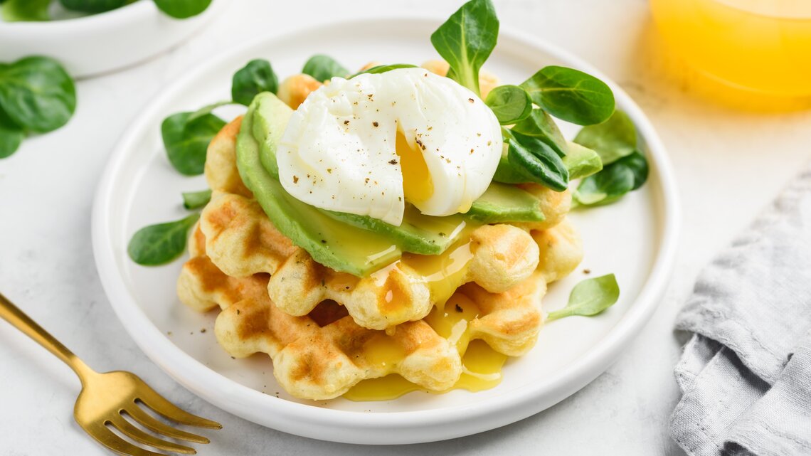 Käsewaffel mit Avocadoscheiben und pochiertem Ei oben, auf einem weißen Teller mit ein paar Feldsalatblättern garniert