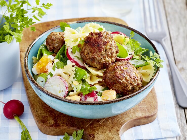 Buletten mit Nudelsalat, bestehend aus gekochten Eiern, Radieschen und Gurken, garniert mit frischem Kerbel und serviert in einer blauben Tonschale auf einem Holzbrett. 