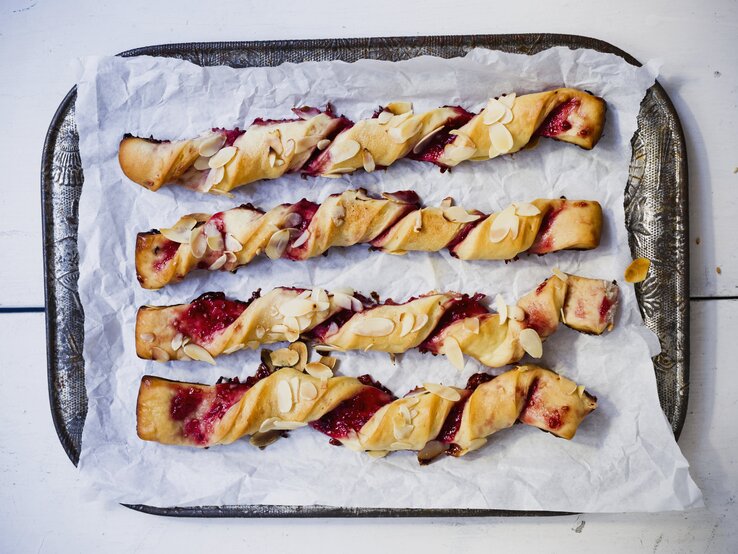 Auf einem Tablett liegen vier Rollen Blätterteigstangen mit Marmelade. Sie sind mit Mandelblättchen verziert.