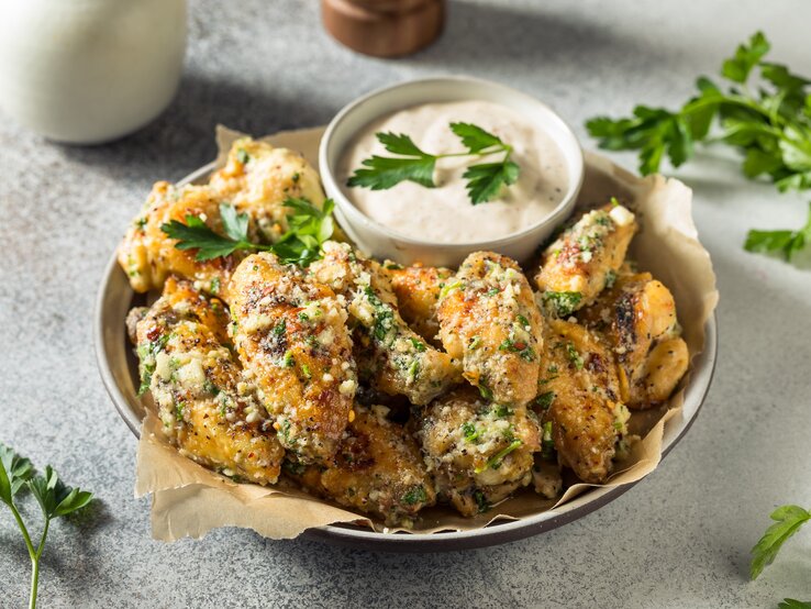 Eine Schale mit gegrillten Parmesan Chicken Wings auf hellem Untergrund mit Petersiliengarnitur und einer Schale hellem Dip. Daneben ein Bund Petersilie. Oben Pfeffermühle. Aufnahme aus der Vogelperspektive.