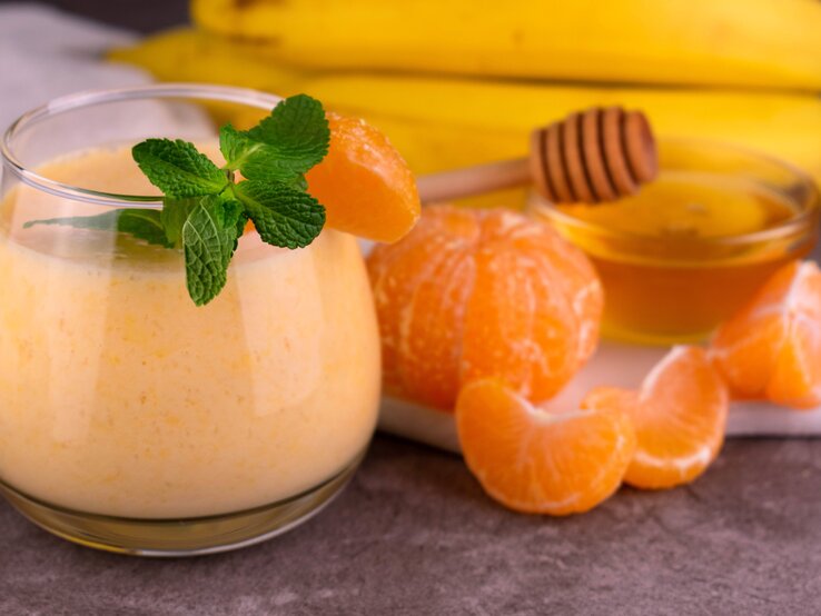 Ein Glas mit dem Smoothie. Daneben Mandarinen und Bananen sowie eine kleine Glasschale mit Honig.
