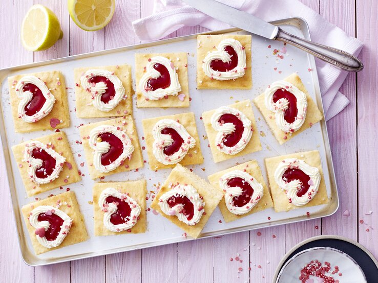 Auf einem Backblech liegen einige Stücken des Zitronen-Herz-Kuchen. Sie sind dekoriert mit herförmiger Mascarpone-creme und Erdbeerkonfitüre. 