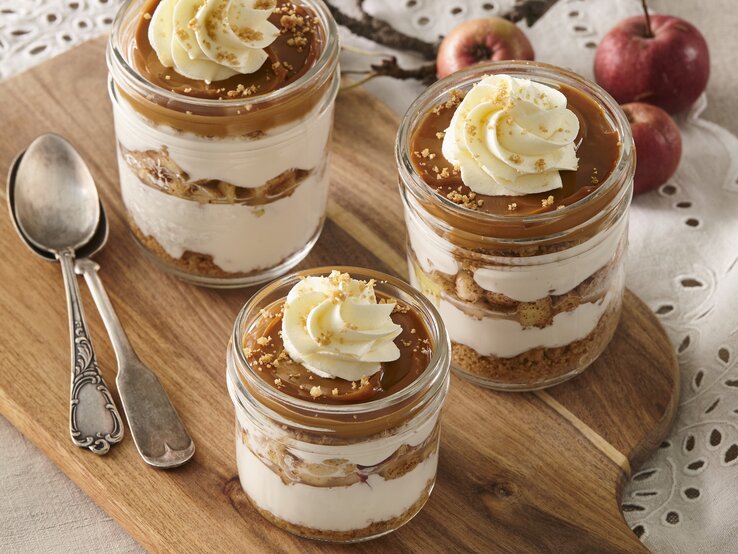 Auf einem Holzbrett stehen vier Gläser Käsekuchen im Glas mit Karamell und Apfel. Sie sind verziert mit Sahne und Keksen. Daneben liegen zwei Löffel. 