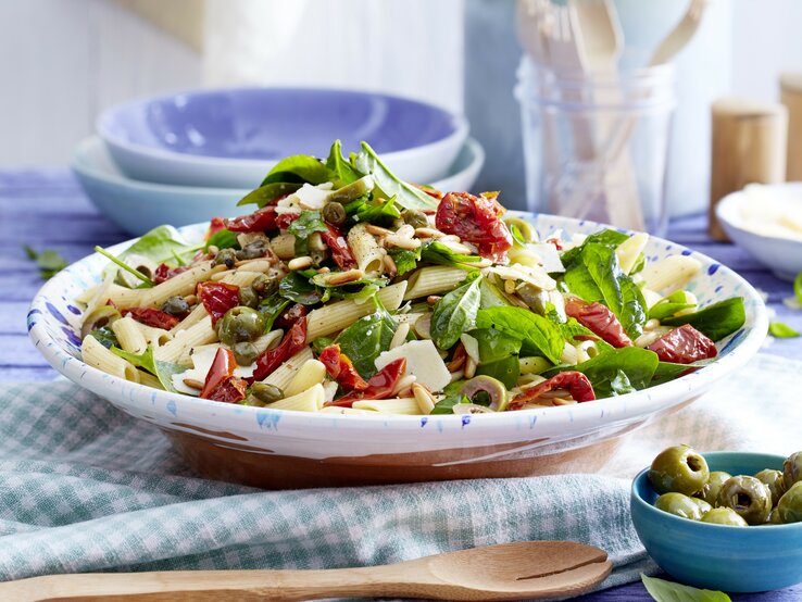 Italienischer Nudelsalat mit Kapern, Oliven, getrockneten Tomaten, Babyspinat und Parmesan in einer hellblauen Schale auf Küchentüchern gereicht.  | © EAT CLUB / Isabell Triemer