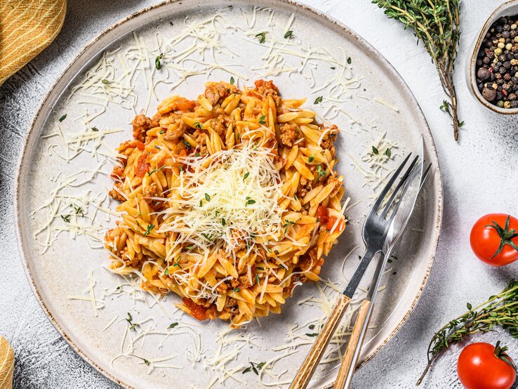 In unsere One-Pot-Orzopizza wandert nicht nur Nudelreis, sondern auch Zucchini, Tomaten und kleine Maiskörner. Ein Teller veganes Orzorisotto in der Draufsicht, mit veganem Parmesan bestreut, daneben Besteckt, ein Geschirrtuch, Tomaten und Gewürze.