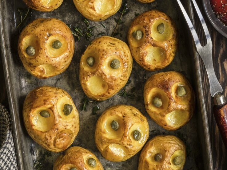 Kreativ, einfach und lecker: Halloween-Bratkartoffeln-mit-Kapernaugen Halloween-Bratkartoffeln mit Kapernaugen, geschnitzt mit gruseligen Fratzen und serviert auf einem schwarzen Blech auf hölzernem Untergrund.