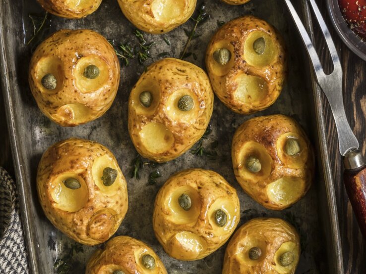 Kreativ, einfach und lecker: Halloween-Bratkartoffeln-mit-Kapernaugen Halloween-Bratkartoffeln mit Kapernaugen, geschnitzt mit gruseligen Fratzen und serviert auf einem schwarzen Blech auf hölzernem Untergrund.