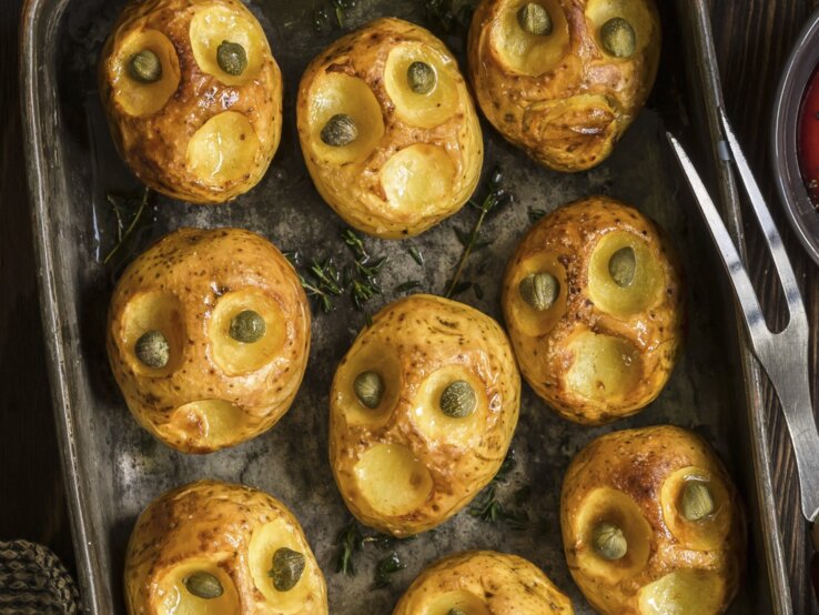 Kreativ, einfach und lecker: Halloween-Bratkartoffeln-mit-Kapernaugen Halloween-Bratkartoffeln mit Kapernaugen, geschnitzt mit gruseligen Fratzen und serviert auf einem schwarzen Blech auf hölzernem Untergrund.