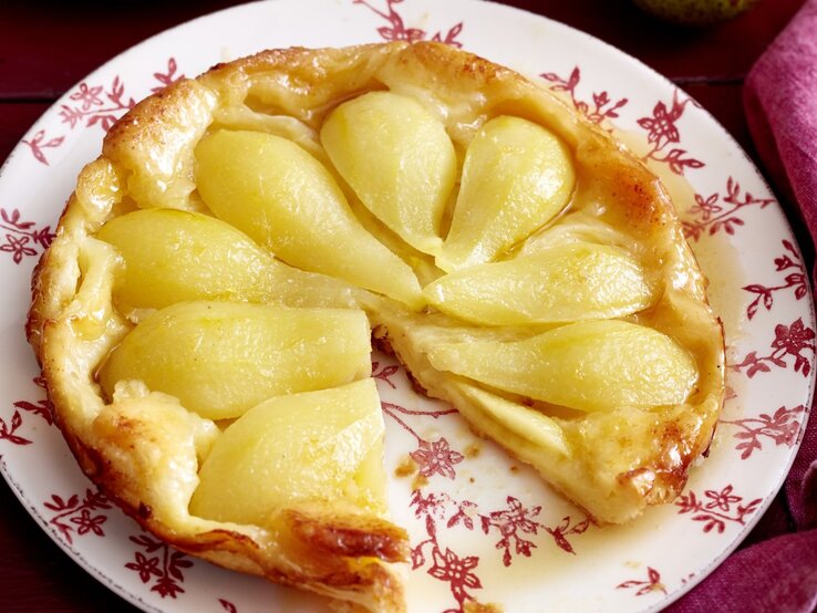 Auf einem Teller liegt eine Birnen-Tarte-Tatin. Sie ist goldbraun gebacken und mit saftigen Birnen belegt.