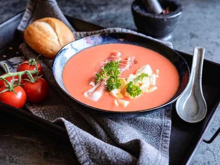 Kalte Tomatensuppe mit gekochtem Ei & Schinken serviert in schwarzer Schale auf schwarzem Tablett. Umgeben ist das Gericht von Tomaten, einem Brötchen und Besteck.