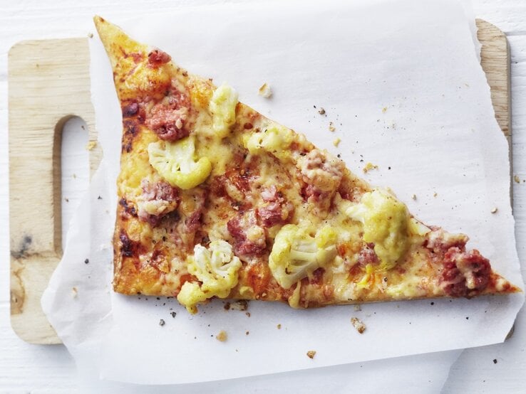 Auf einem Schneidebrett liegt ein Stück Pizza auf norddeutsche Art mit Blumenkohl und Mettwurst. Sie ist frisch gebacken, goldbraun und direkt von oben fotografiert. 