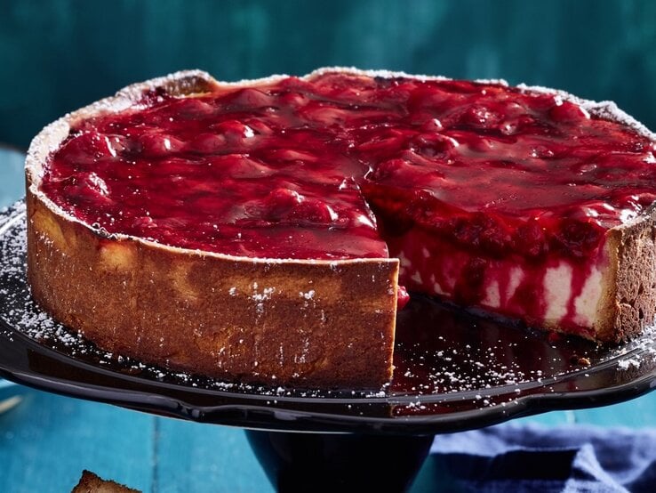 Auf einem Tortenständer steht eine Schmandtorte mit Beeren-Topping. Sie ist angeschnitten und man erkennt die cremige Füllung. 