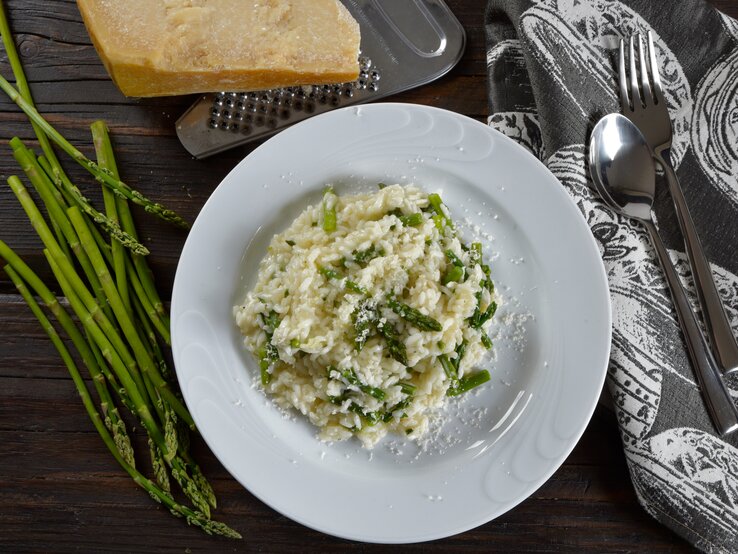 Draufsicht: Eine weißer Teller Wildspargel-Risotto steht auf einem dunklen Untergrund. Drum herum liegen wilder Spargel, Parmesan, ein Reibe, Besteck und ein Tuch.