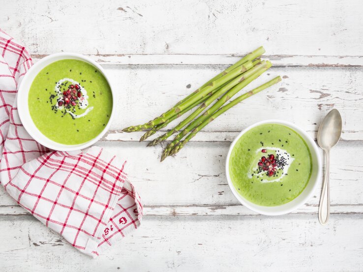 Untergrund aus Holz stehen zwei Schalen. In jeder Schale ist Spargel-Suppe mit Granatapfelkernen und Sesam-Topping / Handtuch und  Löffel / Draufsicht