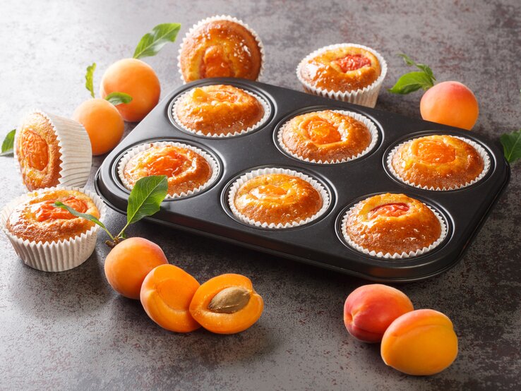 Muffingblech mit Aprikosen-Joghurt-Muffins. Daneben liegen einzelne Muffins und Aprikosen. 