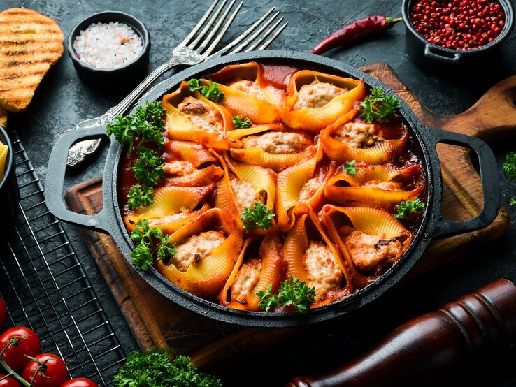 Draufsicht: eine dunkle Pfanne mit gefüllten Muschelnudeln mit Schmand-Hackfleisch steht auf einem Holzbrettchen. Drumherin liegen Schälchen mit Gewürzen, eine pfeffermüfle, Tomaten und rohen Muschelnudeln.