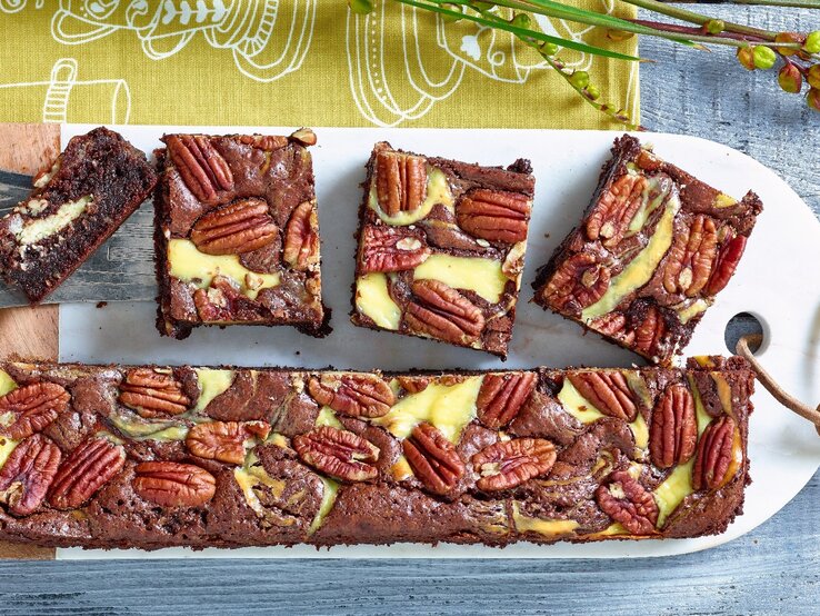Brownies mit Frischkäse und Pekannuss auf weißem Schneidebrett, gelb-grünem Tuch und blauem Tisch. Brownies der Länge nach halbiert. Eine Hälfte zu Rechtecken geschnitten. Das Brett ist gelocht und eine Lederschnur durchgefädelt. Links ein Stück Brwonie auf silbernen Tortenheber mit Intarsien. Oben Zweige.