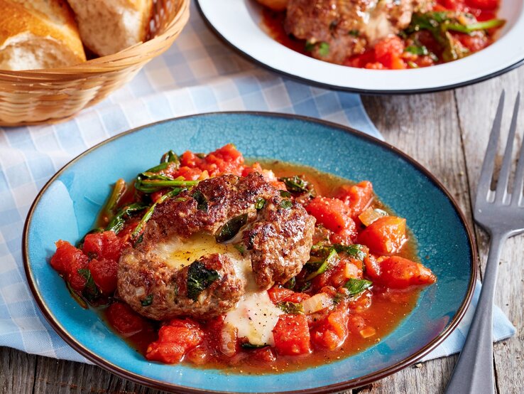 Die Bärlauch-Frikadellen mit dem geschmolzenen Käse auf Gemüseragout. Alles auf einem Blauen Teller und rustikalem Holztisch.