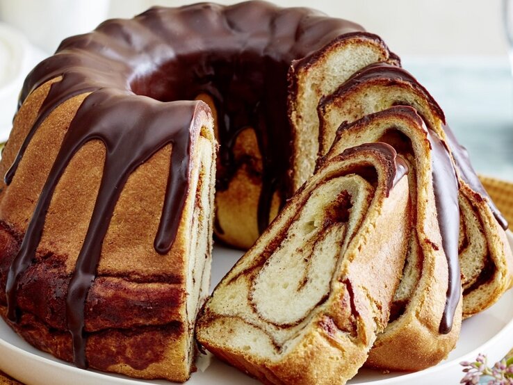 Auf einem Teller steht ein angeschnittener Zimtschnecken-Gugelhupf. Er ist mit einer Schokoladenglasur überzogen. 