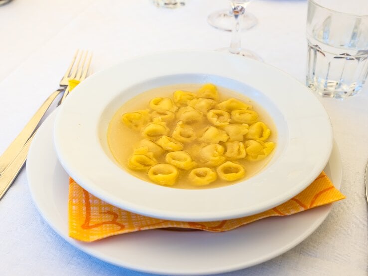 Teller mit klarer Brühe und Tortellini auf weißem Tischtuch. 