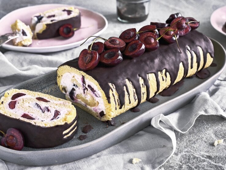 Minibiskuitrolle mit Kirschen und Schokoguss auf einer grauen Servierplatte auf einem grauen Tischtuch neben rosa Tellern auf einem grauen Steintisch. 